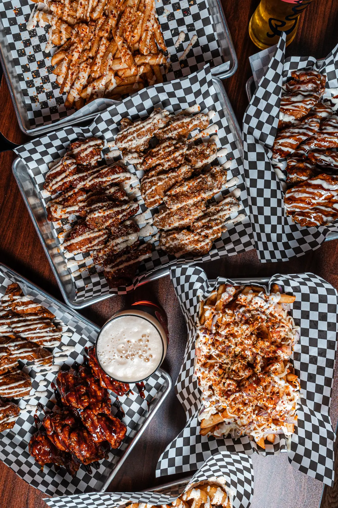 Overhead view of trays of sauced chicken wings, seasoned fries and loaded fries on a wooden table at Sauce N Toss